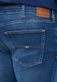 Mörkblå denimjeans med detaljerad sömnad, en bakficka med en liten rektangulär logotyp och en tan läderlogotyp vid midjan.