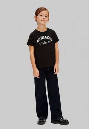 Jeune personne aux cheveux bruns portant un t-shirt noir Calvin Klein, un pantalon large noir et des chaussures noires, debout devant un fond gris.