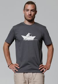 Grijze t-shirt met korte mouwen, voorzien van een witte afbeelding van een papieren boot met een astronaut, tegen een lichtgrijze achtergrond.