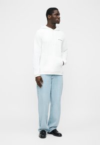 Hombre con sudadera blanca con capucha, vaqueros azul claro y zapatos negros, de pie con una mano en el bolsillo, mirando hacia un lado.