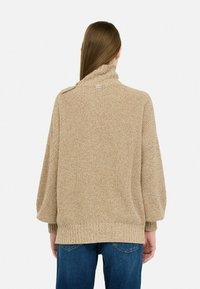 Maglione beige lavorato a maglia con colletto alto, motivo testurizzato e dettagli con bottoni laterali. Abbinato a jeans in denim blu, con una vestibilità comoda.