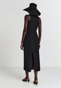 Schwarzes ärmelloses Kleid mit seitlichem Schlitz, aus glattem Stoff gefertigt. Begleitet von einem breitkrempigen schwarzen Hut und offenen schwarzen High Heels.
