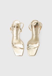 Sandales à talons hauts dorées avec une finition métallique brillante, design à bout carré et fines brides croisées. Semelle plate avec des détails minimaux.