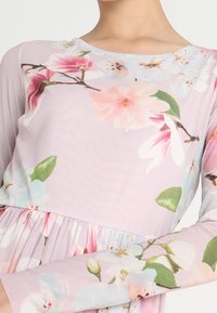 Kukkamekko vaaleanpunaisesta kankaasta. Ominaisuuksina pitkät hihat, pyöreä pääntie ja kukkakuviot vaaleanpunaisina ja sinisinä vihreillä yksityiskohdilla.