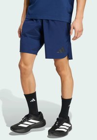 Hombre vestido con pantalones cortos deportivos azul marino, calcetines negros de Adidas y zapatillas negras de Adidas con rayas blancas, de pie contra un fondo sencillo.
