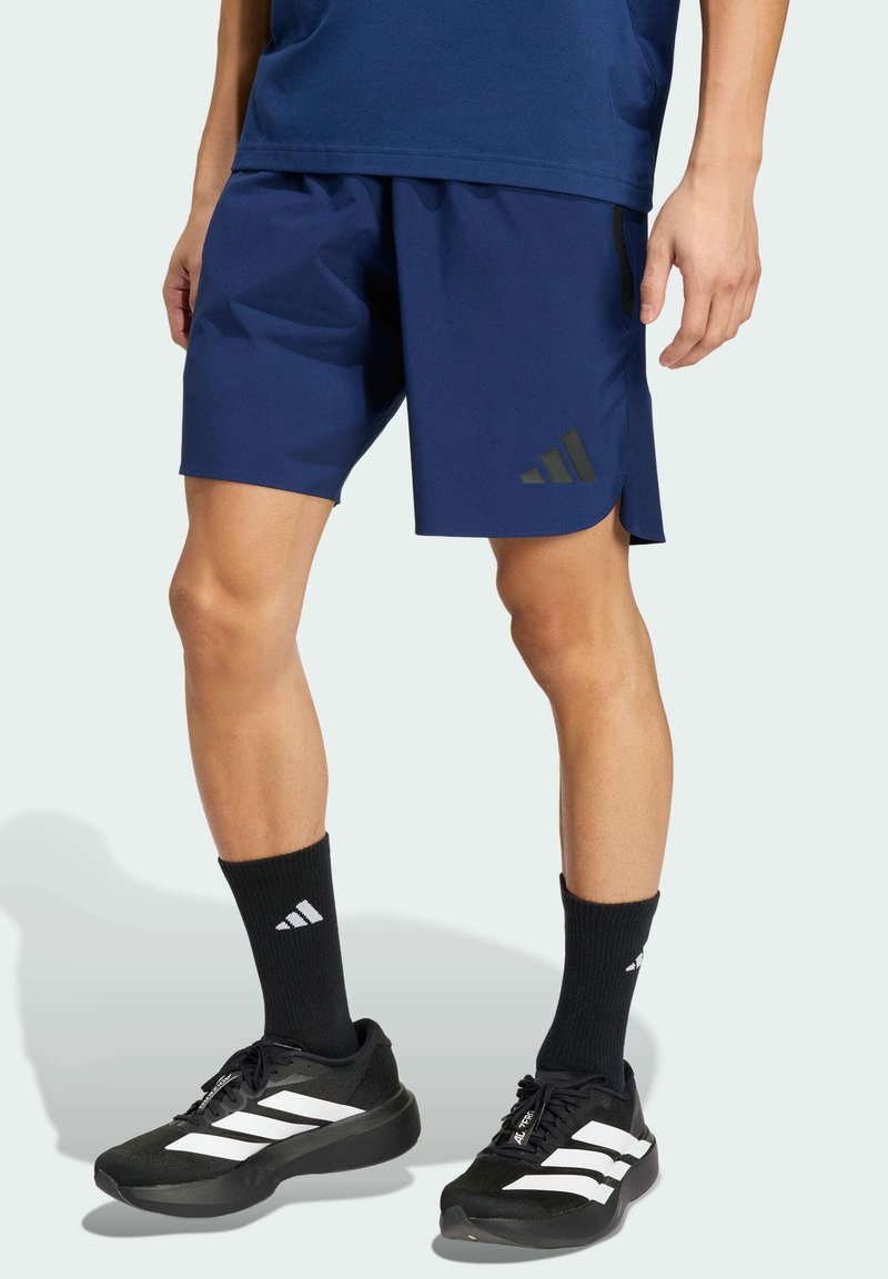 Hombre vestido con pantalones cortos deportivos azul marino, calcetines negros de Adidas y zapatillas negras de Adidas con rayas blancas, de pie contra un fondo sencillo.