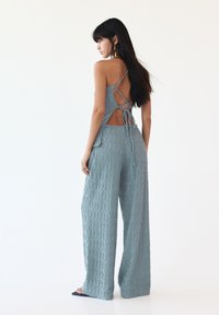 Light blue knit jumpsuit with a textured wavy pattern, open back design with lace-up detailing, and wide-leg trousers.