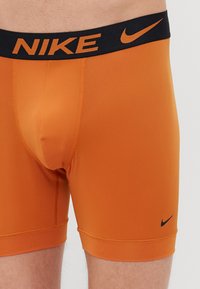 Boxers de nylon laranja com uma cintura elástica preta onde está escrito "NIKE" em letras laranjas. Inclui um pequeno logótipo da Nike em preto na perna.