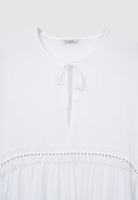 Blouse blanche avec un col rond et un détail à nouer. Présente une bordure en dentelle et de petits accents dorés sur le tissu. Texture légère et lisse.