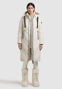 Cappotto lungo imbottito crema con motivo a zigzag, cappuccio, zip laterali e dettagli neri; abbinato a stivali crema coordinati.
