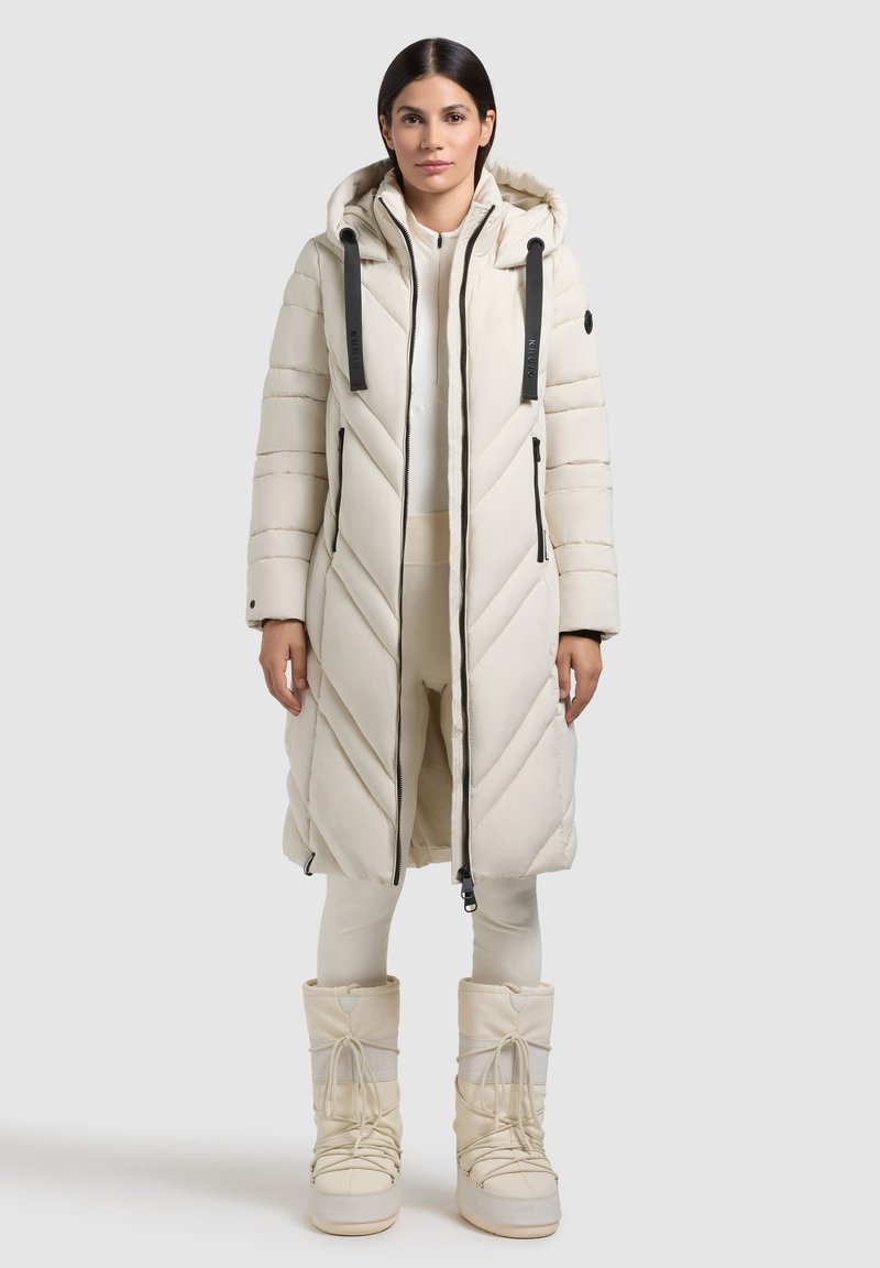 Cappotto lungo imbottito crema con motivo a zigzag, cappuccio, zip laterali e dettagli neri; abbinato a stivali crema coordinati.