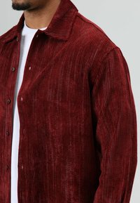 Rode corduroy jas met een getextureerd verticaal streeppatroon, puntkraag, knopen aan de voorkant en twee zichtbare zakken. Draagt over een wit shirt.