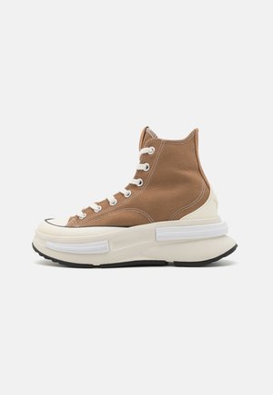 Zapatilla alta marrón con cordones blancos y suela de goma blanca gruesa, suela negra y costuras blancas en la parte superior de lona.