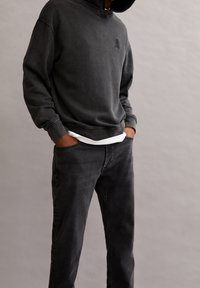 Černá mikina s kapucí s texturovaným povrchem a předním logem, v kombinaci s tmavě šedými džíny se bočními kapsami a pohodlným střihem.