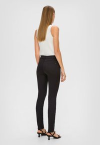 Jean noir slim fit taille haute, avec deux poches arrière et une texture lisse. Porté avec un haut sans manches blanc et des sandales noires.
