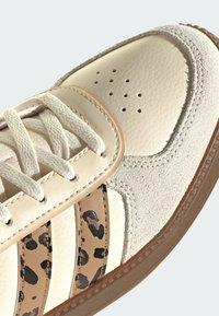 Kremowy skórzany sneaker z zamszowym noskiem, paskami w panterkę, kremowymi sznurowadłami i brązową gumową podeszwą na jasnym tle.