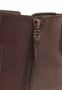 Braune Lederstiefeletten mit strukturiertem Fischgrat-Muster, ausgestattet mit einem seitlichen Reißverschluss und genähten Akzenten. Nahaufnahme des Reißverschlussbereichs.