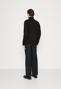 Black cotton sweatshirt with a high collar, paired with loose-fitting black trousers and black shoes, viewed from the back.