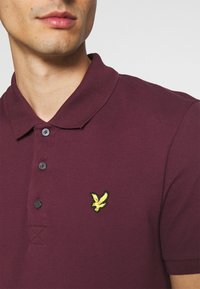 Burgundské polo tričko s límečkem, tříknoflíkovou légou a žlutým logem na levé straně hrudi. Vyrobeno z texturované bavlněné tkaniny.
