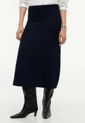 Kvinde iført et sort midinederdel, hvid langærmet skjorte med opviklede ærmer, sorte læderstøvler med hæl og et guld-armbåndsur.