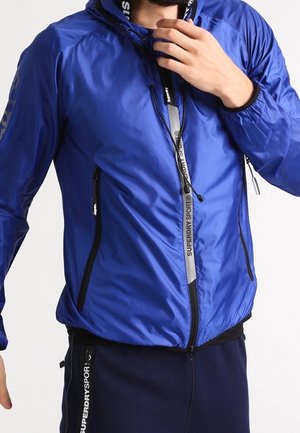 Homme portant une veste zippée bleue Superdry Sport ajustant le col, associée à un pantalon bleu marine Superdry Sport.