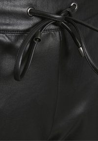 Des pantalons noirs en cuir présentent une texture lisse avec une fermeture à cordon, des accents de quincaillerie en argent et des détails de couture subtils.