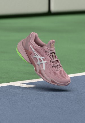 Roosa ja valge ASICS spordijalats laimirohelise tallakujundusega, hõljub tenniseväljakupinna kohal valge piirjoone lähedal.