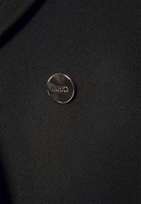 Bouton rond argenté avec l'inscription "LIU•JO", fixé sur un fond en tissu noir texturé.