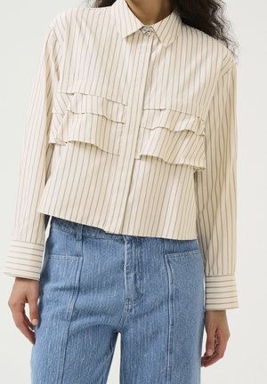 Kobieta ubrana w kremową koszulę z długim rękawem, z pionowymi paskami i warstwowymi falbankami z przodu, w połączeniu z jasnoniebieskimi jeansami z wysokim stanem.