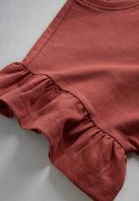 Detailný záber na červenohnedú látku s volánovým švom a viditeľným prešívaním na okraji odevu.