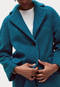 Abrigo de lana texturizada en color azul verdoso con cuello con muesca, grandes bolsillos y cierre de un solo botón. Llevado sobre un suéter blanco acanalado.