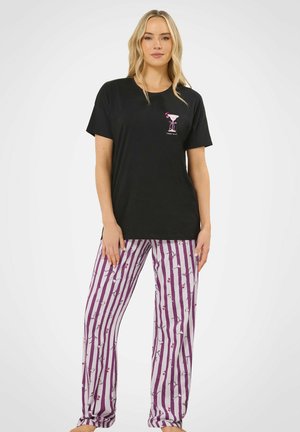 Donna che indossa una maglietta nera con la grafica di un bicchiere da cocktail e pantaloni del pigiama a righe viola e bianche con stampa di ciliegie, in piedi a piedi nudi.