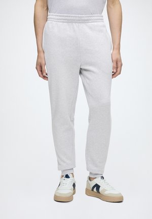 Partie inférieure du corps d'une personne portant un pantalon de survêtement gris clair et des baskets blanches avec des accents bleu foncé, debout sur un fond blanc uni.