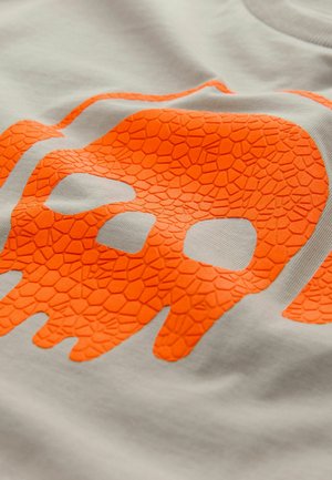 Világosszürke póló, középen elhelyezett, kiemelkedő, textúrával ellátott narancssárga koponya grafikával, amely repedezett mintával díszített.