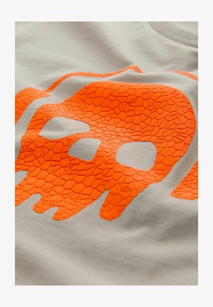 Svijetlosiva majica s teksturiranim narančastim grafikonom lubanje koja ima pukotinasti uzorak, smještena središnje i istaknuta.