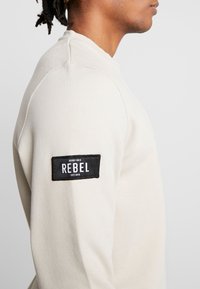 Sweat-shirt beige clair avec un col arrondi et des poignets côtelés. L'épaule gauche porte un patch rectangulaire noir affichant "REBEL" en lettres blanches.