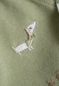 Un tessuto verde presenta un dachshund ricamato in bianco e marrone che indossa una cravatta verde, posto su uno sfondo strutturato.
