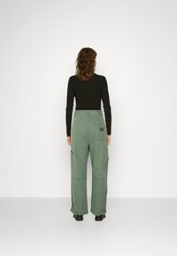 Calças de carga verdes com um corte descontraído, múltiplos bolsos laterais e detalhes em costura visíveis, combinadas com uma blusa preta de manga longa.