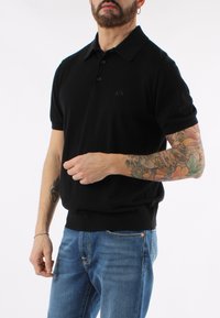 Svart pikétröja med klassisk krage och korta ärmar, prydd med en diskret logo på bröstet, kombinerad med blå denimjeans.