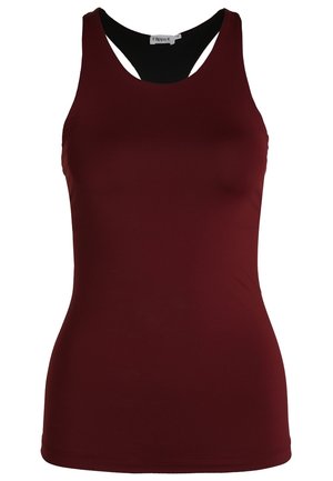 Henkilö istuu lattialla jalat ristissä, päällä hihaton viininpunainen toppi ja tummat legginsit, kädet pään yläpuolella.