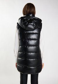 Schwarze Steppweste mit Kapuze, glänzender Oberfläche, genähtem Design und leicht verlängertem Schnitt, getragen über einem weißen Hemd.