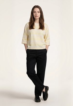 Žena stojí so zdvihnutými rukami v vreckách, oblečená v svetlo žltej pruhovanej košeli, čiernych nohaviciach a čiernych topánkach na jednoduchom pozadí.