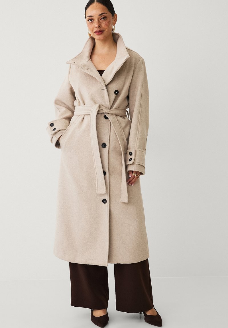 Beige wollen jas met een brede kraag, dubbelrijige ontwerp, zwarte knopen en een bijpassende riem. Gepaard met bruine wijde broek.