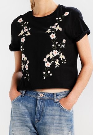 Czarny krótki t-shirt z różowym i białym kwiatowym haftem noszony z jasnoniebieskimi, wysokim stanem jeansami, ręce w kieszeniach.