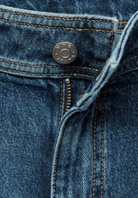 Donkerblauwe denim jeans met gouden stiksels. Voorzien van een metalen knoop met de tekst "MNG JEANS" en een ritsluiting. Close-up detail.
