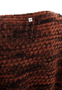 Un tissu tricoté brun et noir avec une surface texturée présente un motif de petites sections en relief. Une étiquette de marque argentée est visible.