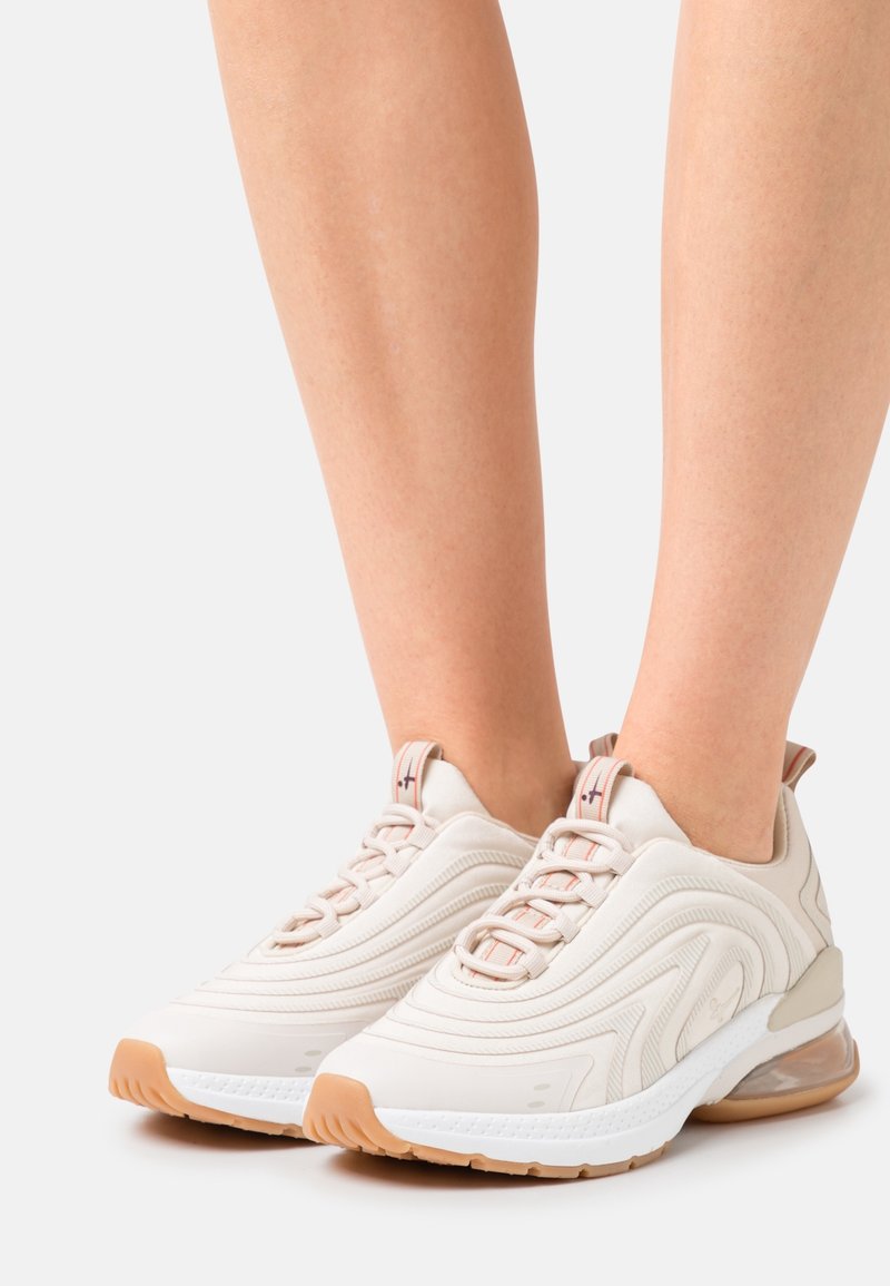 Beige athletic shoes with textured upper, thin, wavy designs, and a cushioned sole. Rubber outsole in light brown with clear air bubble accents.