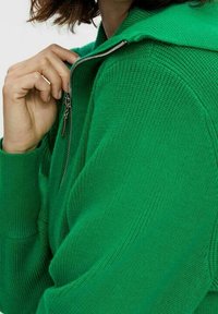 Pull en tricot vert avec capuche, doté d'une fermeture à glissière. Le tissu présente un motif texturé et les poignets sont côtelés.