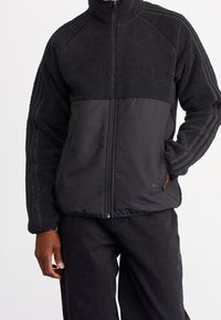 Black jacket featuring a fleece upper and smooth lower panel, zip front, high collar, and textured surface with side stripes.