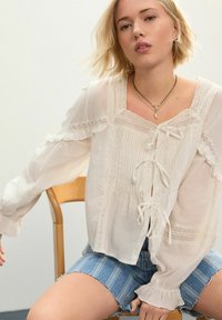 Blouse en coton blanc avec des détails en dentelle, manches bouffantes et liens au centre. Portée sur des shorts en denim bleu avec des bords effilochés.